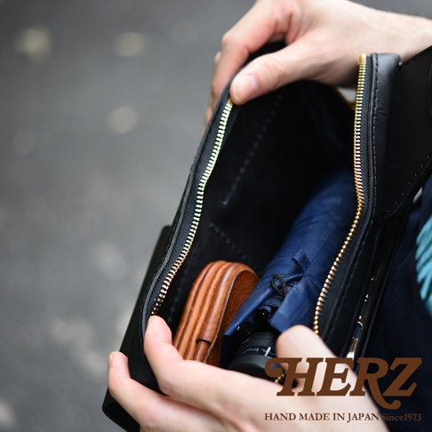 職人手作皮革雙口袋斜背包，黑色皮革材質，內部裝有藍色摺疊傘和棕色皮革製品，金色拉鍊。