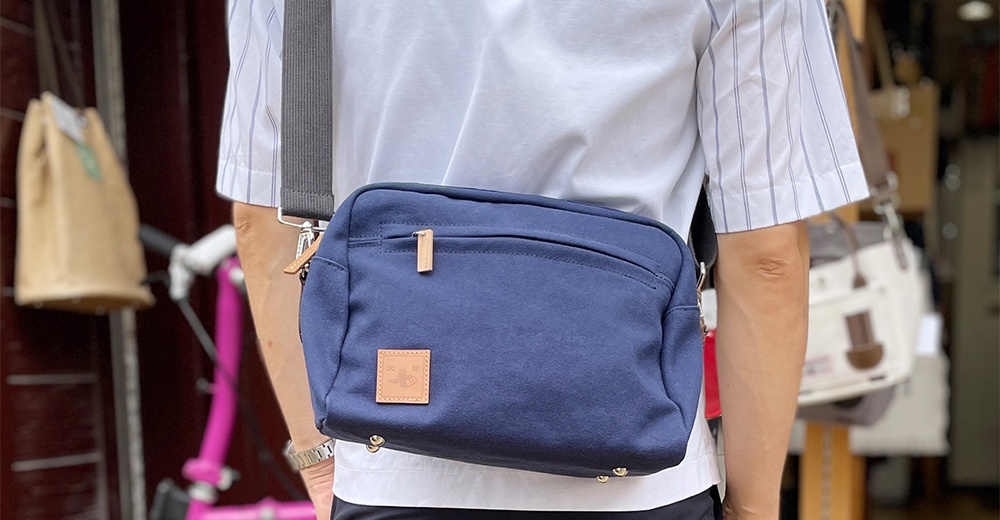 深藍色石洗帆布側背包，配有木質拉鍊頭和方形皮質標籤，由一位身穿條紋襯衫的男性背在肩上