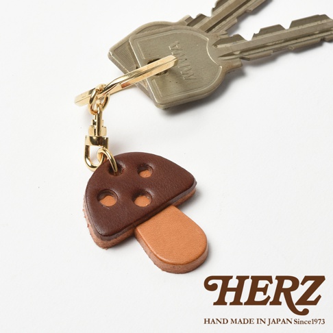 職人手作皮革香菇造型鑰匙圈，深棕色菌蓋點綴四個淺棕色圓孔，搭配淺棕色菌柄，掛有金色鑰匙圈與鉤扣，下方為品牌標誌。
