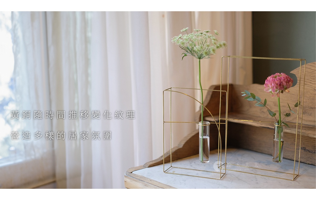 黃銅書架款掛擺兩用花器，以簡約線條勾勒出幾何框架，內含玻璃試管，分別插入白色傘形小花和粉色圓形花朵，展現黃銅質感與自然花藝的結合。