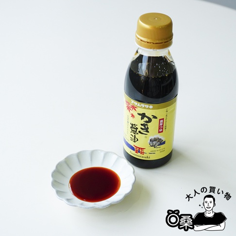 一瓶瓶身標示著朝紫廣島牡蠣醬油的調味料放置在白色桌面上，金色瓶蓋與透明瓶身內裝著深色醬油，旁邊的白色花瓣形小碟子中盛裝著少量醬油，右下角配有動漫風格的人物插圖。