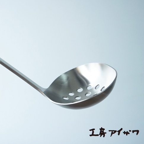 銀色不鏽鋼材質的萬用瀝水湯勺，勺面有圓形孔洞，勺柄細長，置於淺藍色背景中。