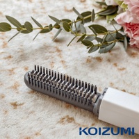 灰色的迷你瀏海梳，梳齒密集，手把為白色，背景有綠葉和粉色花朵裝飾，右下角有KOIZUMI品牌標誌