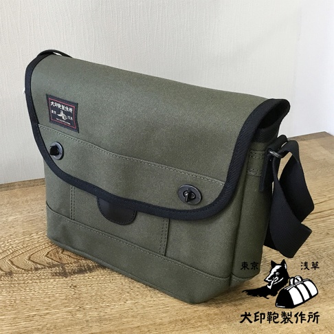 軍綠色純棉帆布製成的大款雙扣郵差包，配有黑色滾邊、黑色皮質裝飾和黑色織帶肩帶。包蓋上有一個黑色標籤，上面印有日文文字和圖案。