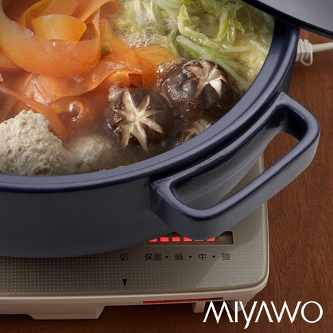 海軍藍陶土湯鍋內燉煮著香菇、貢丸、紅蘿蔔片與白菜，鍋身呈現光滑質地，配有兩側深藍色把手，置於電磁爐上，爐面顯示加熱指示燈。