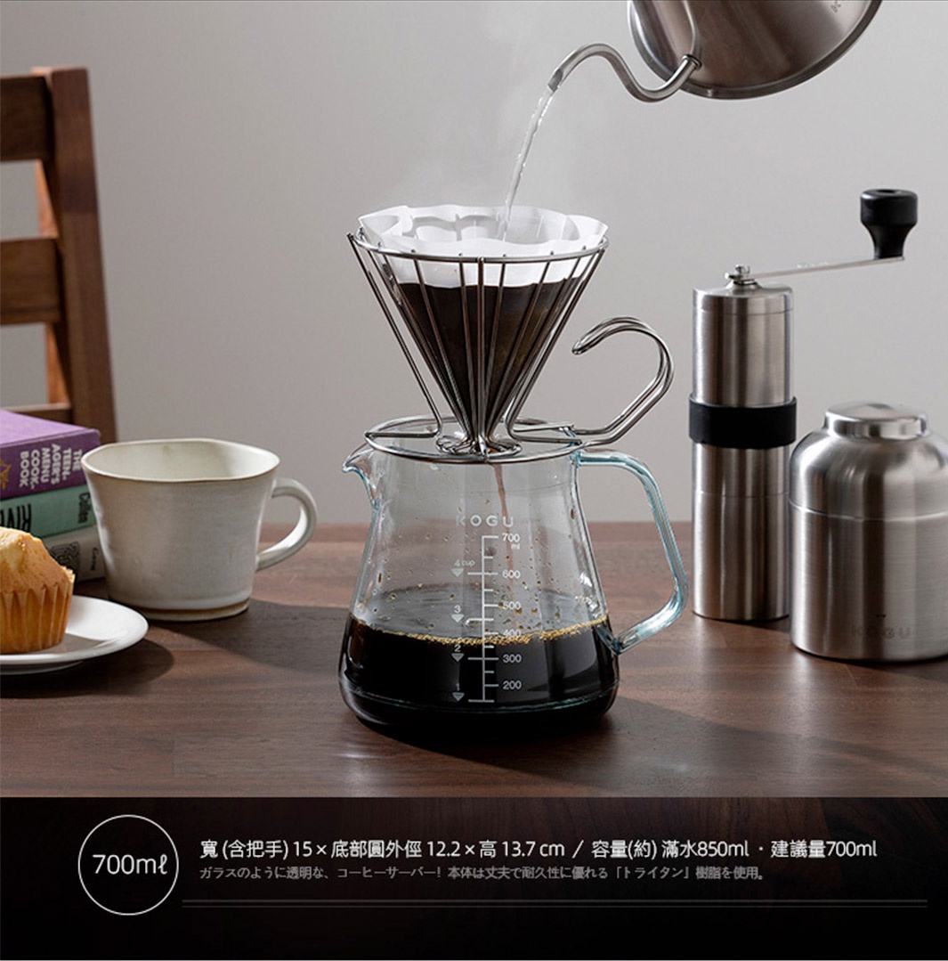 透明玻璃咖啡壺，內有咖啡液，上方為金屬濾網架，內有濾紙和咖啡粉。壺身帶有刻度，容量標示至 700ml。背景有馬克杯、咖啡研磨機及銀色容器。