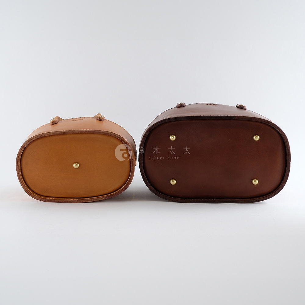 兩個不同尺寸的職人手作皮革束口手提包，分別展示了淺棕色和深棕色的皮革，底部均有金屬腳釘，展現了包袋的結構與質感。