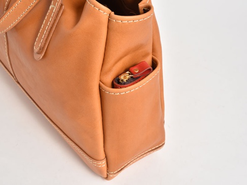 職人手作皮革橫式肩背托特包，採用棕褐色皮革製成，包身車縫線清晰。包側有一個小口袋，內有紅色皮革拉環的拉鍊零錢包。