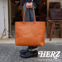 一只手提著一個橘棕色皮革托特包，包包的正面光滑，有著簡潔的縫線。包包上方有兩個手提帶，包包的主體呈現方形。圖片右側有一個深棕色的皮革包，放在一個木製凳子上。圖片下方有HERZ的白色字體標誌。