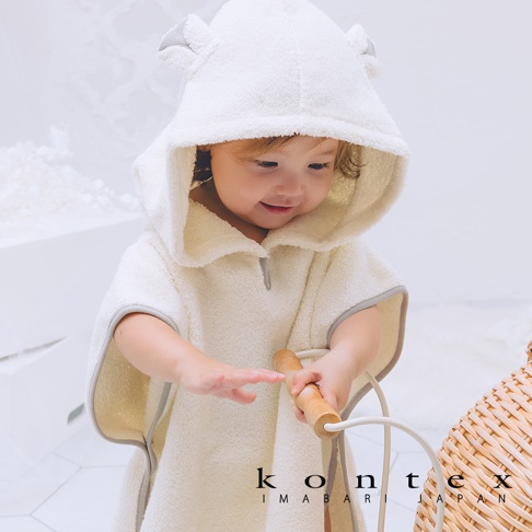 嬰兒穿著奶油色絨毛材質的斗篷式連帽浴巾，帽子上有可愛的熊耳朵造型，浴巾邊緣有淺灰色滾邊，整體溫暖舒適。