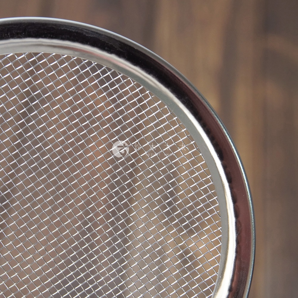 不鏽鋼萬用撈油勺的特寫，展示了細密的銀色金屬網篩和光滑的銀色不鏽鋼邊緣，背景為模糊的木紋。
