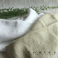 兩條紗布巾，一條為白色，一條為淺褐色，皆呈現柔軟紋理，背景為淺色布料，一旁點綴綠色植物枝葉。