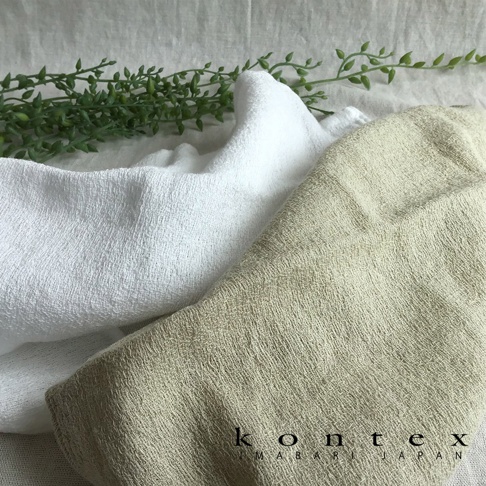 兩條紗布巾，一條為白色，一條為淺褐色，皆呈現柔軟紋理，背景為淺色布料，一旁點綴綠色植物枝葉。