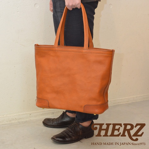 HERZ 手工製作的皮革托特包，採用溫暖的橘棕色皮革，搭配黑色長褲和棕色皮鞋，展現職人質感。