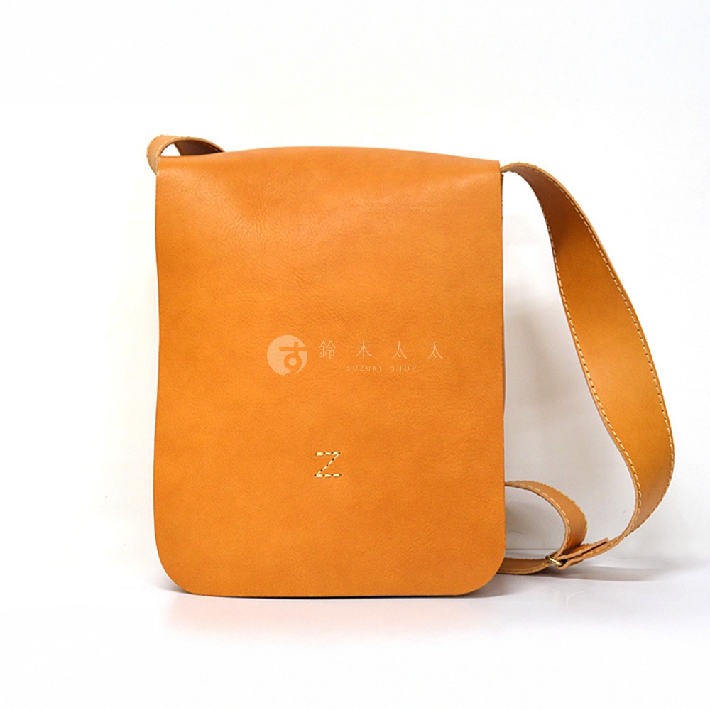 日本HERZ職人手作皮革軟皮直式斜背包，展示一個橘棕色皮革斜背包，包包上有日文「鈴木太太」和英文「SUZUKI SHOP」的品牌標誌，以及一個縫線的「Z」字樣。