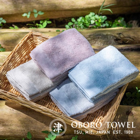 藤編籃子裡擺放著四條不同顏色的毛巾：淺紫、淺灰、淺藍及另一條淺灰。毛巾質地柔軟，邊緣有細緻的條紋。