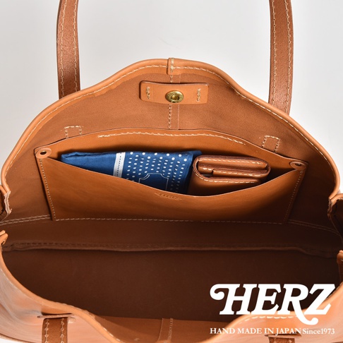 這是一款日本HERZ手工製作的皮革肩背托特包內部俯視圖，呈現棕褐色優質皮革質感，內部設有皮革隔層，放置了一塊藍色圓點布巾與皮革錢包，包口處飾有金屬按扣，下方印有HERZ品牌標誌與手工製造說明。