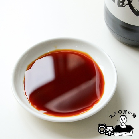 白色小碟中盛有深褐色醬油，醬油表面光滑，有反光。碟子旁有一瓶醬油的部分瓶身，瓶身為黑色與白色，上有日文文字。