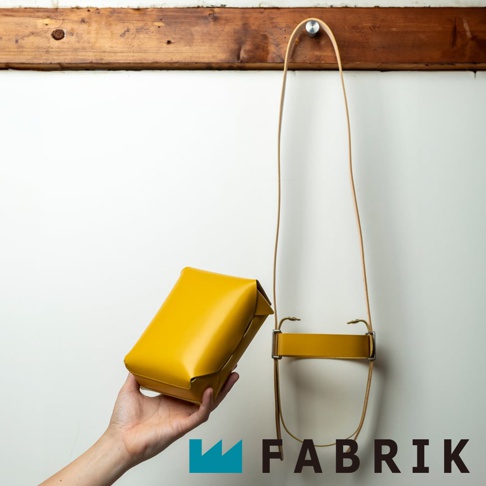 FABRIK 品牌一個亮黃色皮革迷你斜背包，懸掛於牆上的木製橫桿，以及一隻手正拿著該包。