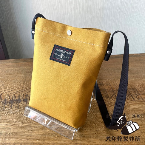 芥黃色帆布製成的方形側背包，包身正面中央有黑色方形布標，印有日文及英文品牌名稱與圖案，黑色背帶從包包上方兩側延伸下來。