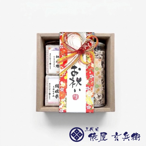 一份精緻的三代目俵屋玄兵衛日本健康米禮盒，包裝盒內整齊排列兩包真空包裝米與一瓶穀物。盒裝中央飾有華麗的紅底花卉圖案腰封，並以日本傳統水引結裝飾，腰封上方以書法字體寫著祝賀詞。整體呈現雅緻的日式伴手禮風格，設計細膩且充滿儀式感。