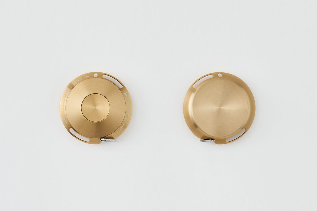 兩個金色鋁製卷尺，一個正面顯示中心圓形結構與周圍刻字，另一個背面顯示光滑的圓形表面，皆配有銀色金屬拉環。