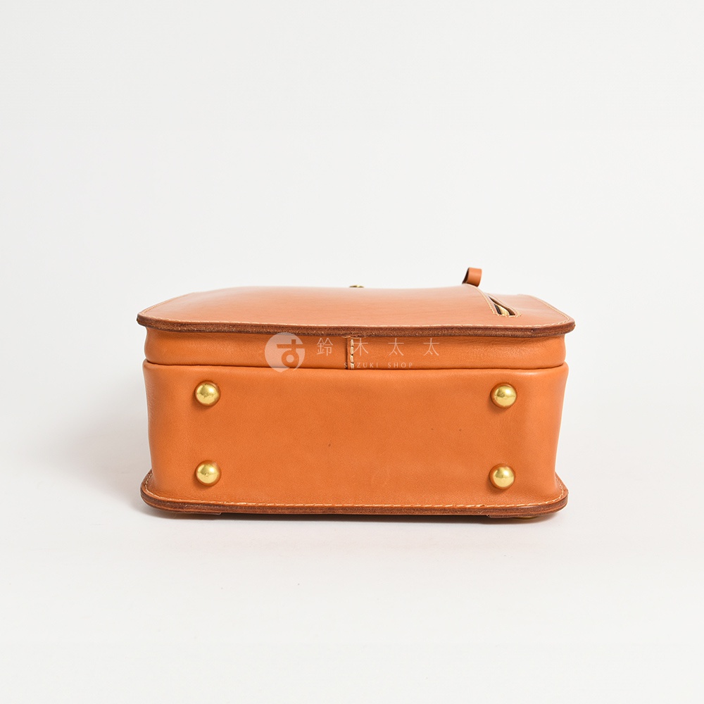 職人手作皮革後背包，採用棕褐色牛皮，方形箱型設計，底部有四個金色銅釘，頂部有皮革提把和拉鍊開口。