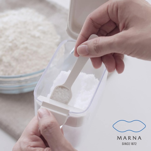 一隻手正使用專屬的小量匙，從透明的Marna密封防潮調味料盒中取出一勺調味料，調味料盒設計有貼心的刮板裝置，可以精準測量並平整勺內的份量，整體呈現簡約清新的居家收納風格。