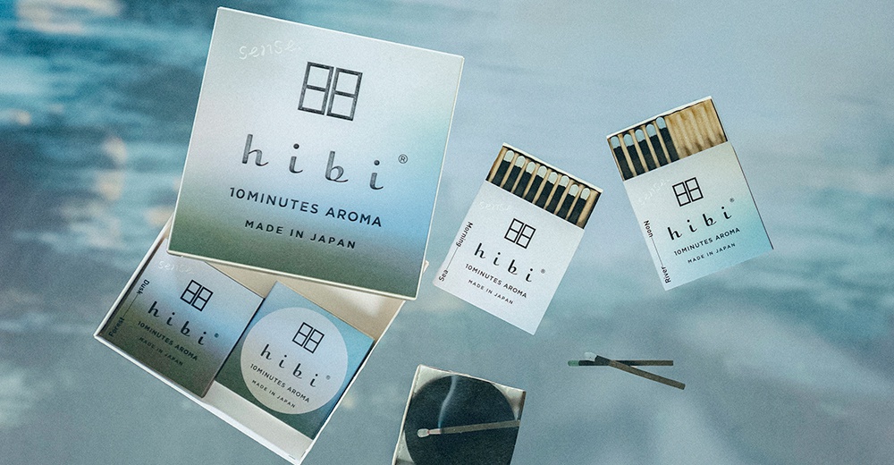 在波光粼粼的水面背景上，漂浮著 hibi 香氛火柴禮盒及其內容物。畫面中央是一個大型禮盒，周圍環繞著標示為 Morning、Sea、Noon、River、Dusk 與 Forest 不同情境的香氛火柴包裝。下方展示了打開的禮盒與正在燃燒的香氛火柴，整體色調呈現優雅清新的質感，展現出 hibi 十分鐘香氛的生活美學。