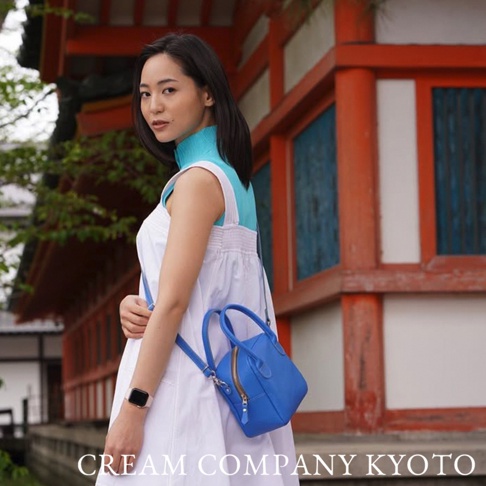 身著白色連身裙的女性，斜挎著一個亮藍色的皮革貝殼包，包身為方形，設有提把和拉鍊。