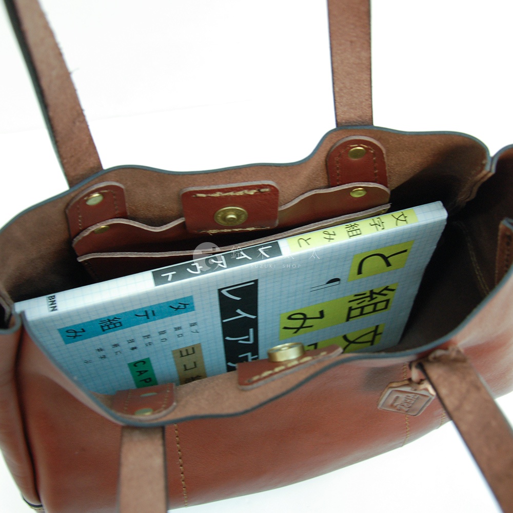 棕色皮革仕女托特包，內部可見隔層與一本日文書籍，包身材質為皮革，顏色為深棕色，配有雙提把。