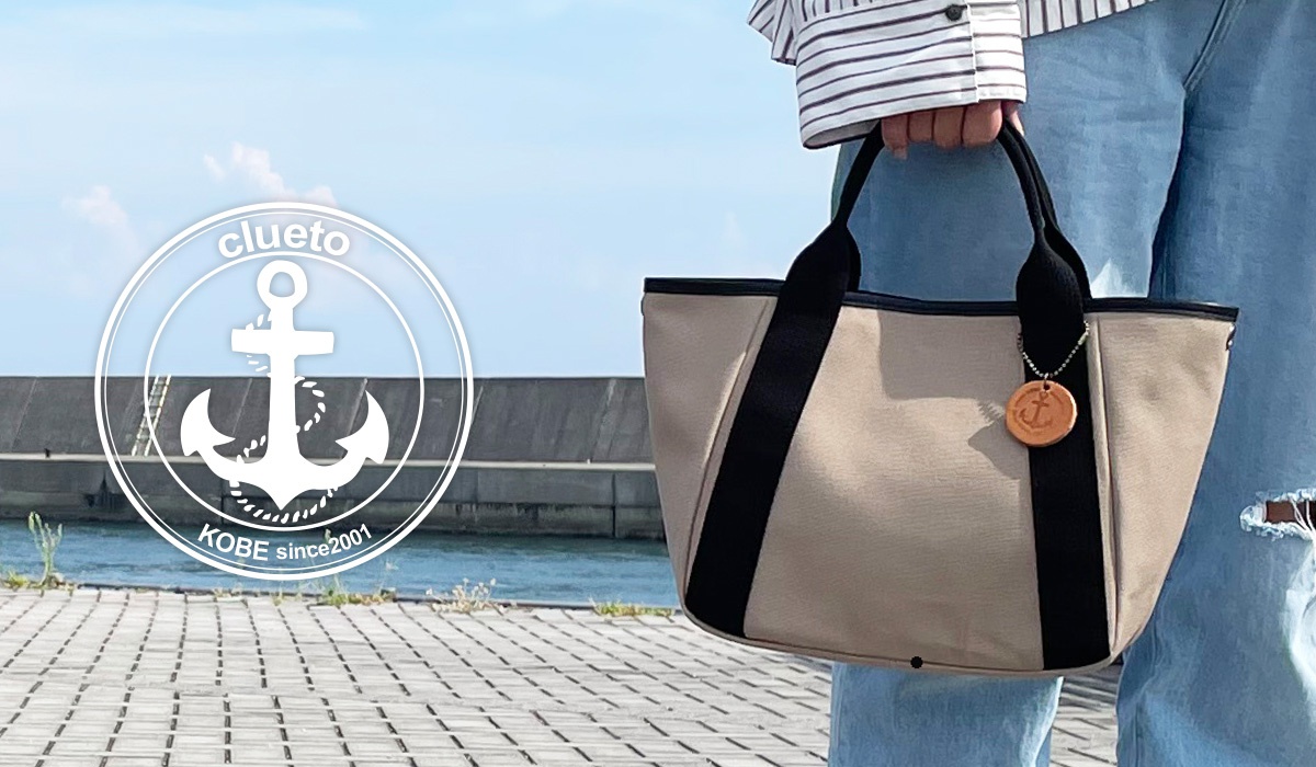 一個人在海邊提著米色帆布托特包，包上掛著船錨吊飾，背景為藍天和海港。