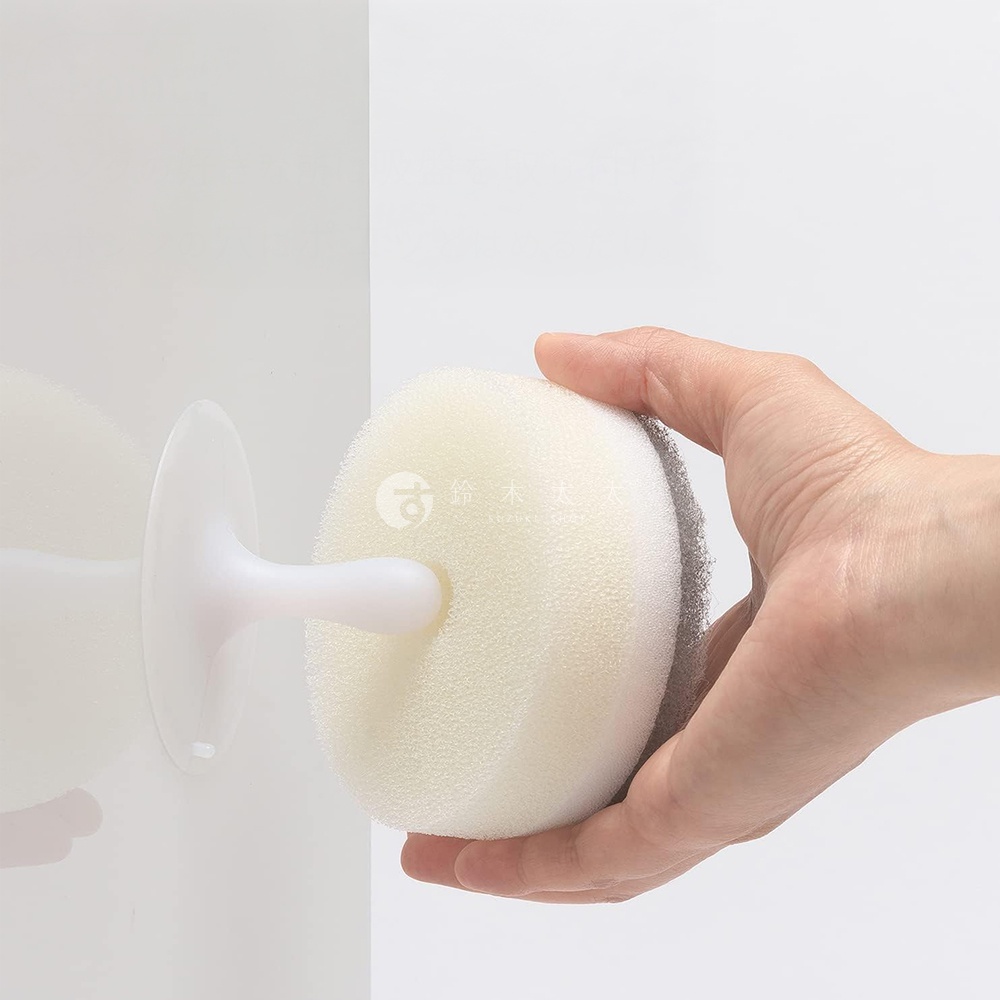 手持圓形白色海綿，背面有灰色研磨層，海綿上印有日文標誌，旁邊有白色吸盤支架黏在白色牆壁上