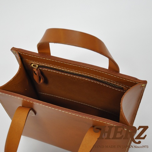 職人手作皮革直式托特包，呈現焦糖棕色，以真皮材質製作，包身縫線清晰，內有拉鍊內袋，提把與包體連接處有鉚釘固定。