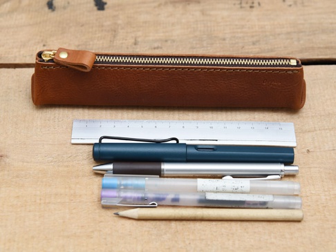 職人手作皮革Organ三角收納筆袋，咖啡色皮革，配有金色拉鍊及皮革拉環。筆袋下方放置一把金屬尺，以及一支深藍色鋼筆、一支銀色原子筆、兩支透明筆桿的筆，還有一支淺棕色鉛筆。