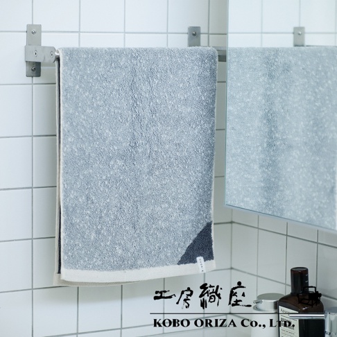 一條淺灰色棉麻混紡毛巾，底部有米白色滾邊，右下角有深藍色幾何圖案。毛巾掛在銀色金屬毛巾架上，背景為白色磁磚牆。