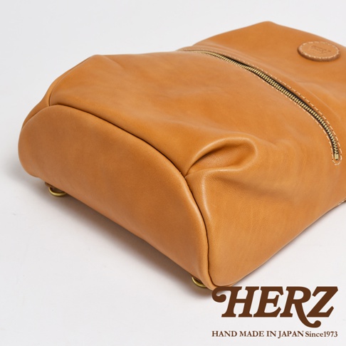 棕色皮革製成的大開口圓角後背包，帶有金色拉鍊，底部有金屬鉚釘，側面有品牌標誌。