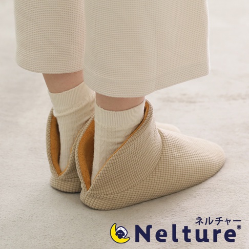 一雙米色菱格紋布面、黃色內襯的保暖腳踝室內拖鞋，由穿著米色寬褲和米色襪子的人穿著，置於淺灰色地毯上。