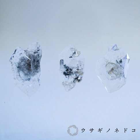 三塊透明水晶，內含黑色雜質，呈現片狀標本特徵，置於淺藍色背景上。