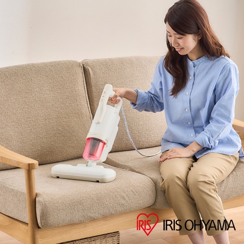 白色手持式除塵蹣機，配備透明粉紅色集塵筒，機身有操作按鈕，連接白色電源線，底部有寬扁吸頭，放置在米色布藝沙發上。