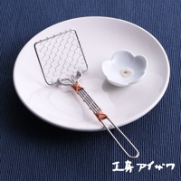 銀色不鏽鋼製成的方形豆腐杓，帶有細網狀勺面，銅色線條纏繞的握柄。旁邊有一個白色小碟，碟上有花朵圖案。