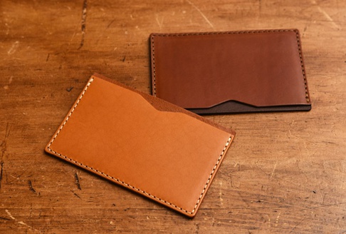 兩件職人手作皮革文件收納夾，一件為淺棕色皮革，另一件為深棕色皮革，皆以白色縫線手工縫製，置於木紋桌面上。