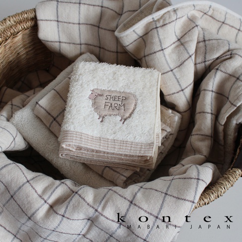今治純棉手帕，白色為主，繡有可愛的綿羊圖案及「SHEEP FARM」字樣，邊緣有細條紋裝飾。多條紋格紋的毛巾圍繞，整體為米白色與褐色調。