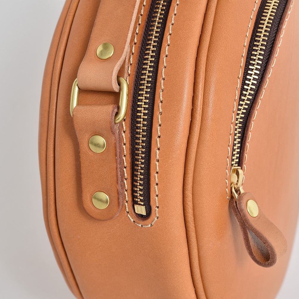 職人手作的淺棕色皮革小圓包，特寫鏡頭展示了兩個黃銅色拉鍊、皮革拉環和黃銅釦，以及皮革的紋理與車縫線。