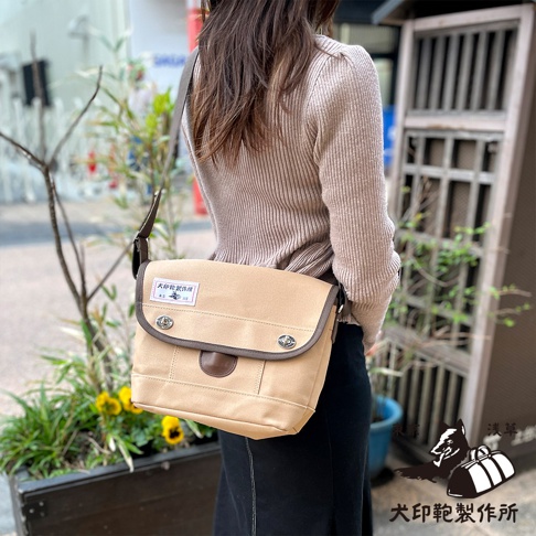 犬印純綿帆布雙扣郵差包，淺卡其色，配有深棕色皮質飾邊和兩個銀色圓形金屬扣，側背於一位身著米色針織衫的女性身上。