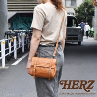 一位女士身著淺駝色上衣和灰色長褲，斜挎一個棕色皮革復古郵差包。包包有翻蓋設計，配有兩個皮帶扣和皮質肩帶。