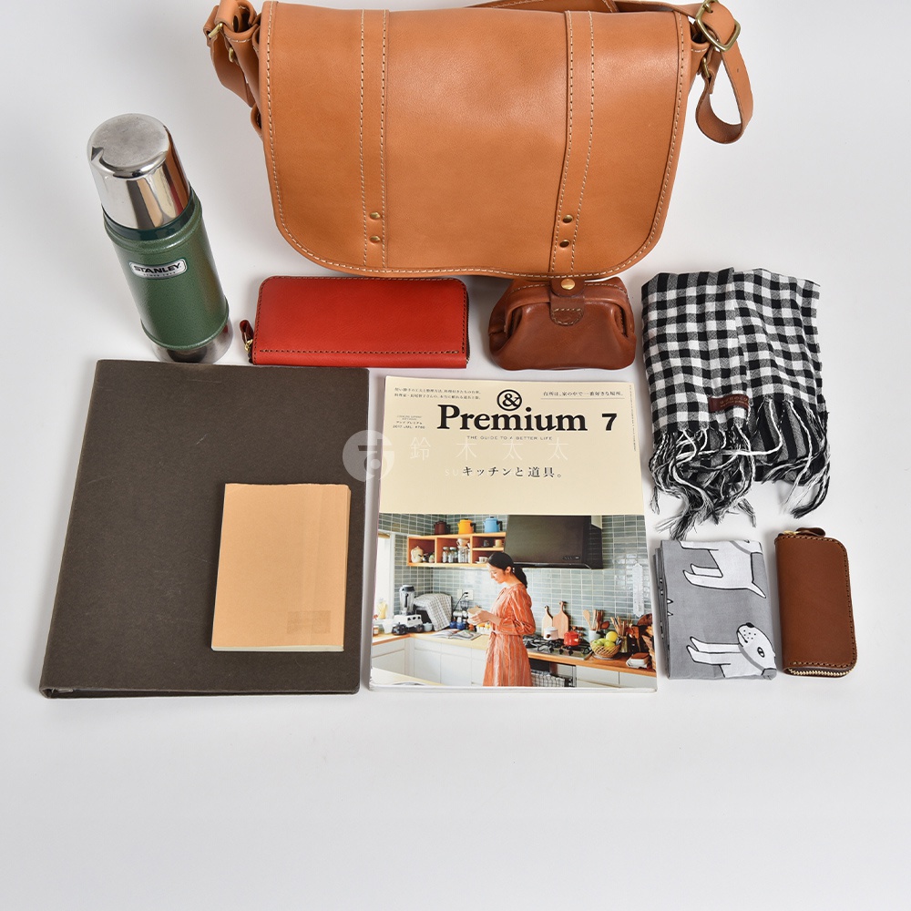 職人手作皮革復古郵差包，淺棕色皮革，縫線整齊。包旁有綠色保溫瓶、紅色皮夾、棕色零錢包、黑白格紋圍巾、灰色印花布料、棕色鑰匙包，以及一本雜誌和一個深灰色筆記本。