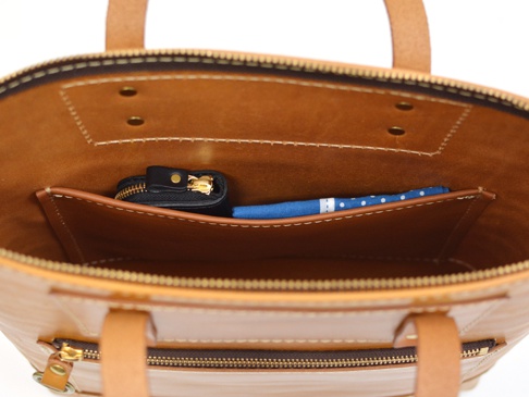 職人手作皮革威靈頓直式拉鍊托特包內部，展示一個黑色拉鍊零錢包和一個藍色印花方巾，置於皮革內袋中。包包主體為淺棕色皮革，配有黃銅色拉鍊及提把。