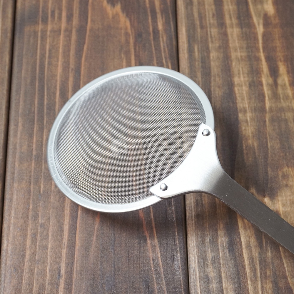 不鏽鋼萬用撈泡勺，圓形細網篩，搭配金屬手柄，整體呈銀色，放置於深棕色木紋桌面。