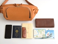 職人手作皮革拉鍊旅行斜背包，採用天然棕色皮革，設有雙拉鍊前袋和金色拉鍊。旁邊擺放著黑色手機、藍色日本護照、棕色皮革鑰匙包、黃色書籍、棕色皮夾、羅馬地圖及國泰航空機票。